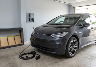 Grey electric hatchback with a dented front wing in a segregated specialist workshop assessment bay, beside closed equipment and green-accented cabinets.